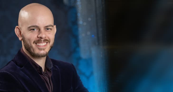 A picture of me at my graduation. I'm wearing a navy blue blazer and a dark purple shirt. In the background, there's a light blue panel with some bright lights. I'm smiling, sporting a short beard, and I'm bald.